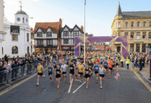 Mass start of a marathon in Stratford-upon-Avon. Runners fill Bridge Street, passing historic half-timbered Tudor buildings on the left, with the stone arches of Clopton Bridge crossing the river in the far distance under a blue sky with clouds.