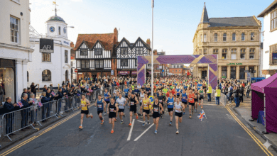 Mass start of a marathon in Stratford-upon-Avon. Runners fill Bridge Street, passing historic half-timbered Tudor buildings on the left, with the stone arches of Clopton Bridge crossing the river in the far distance under a blue sky with clouds.